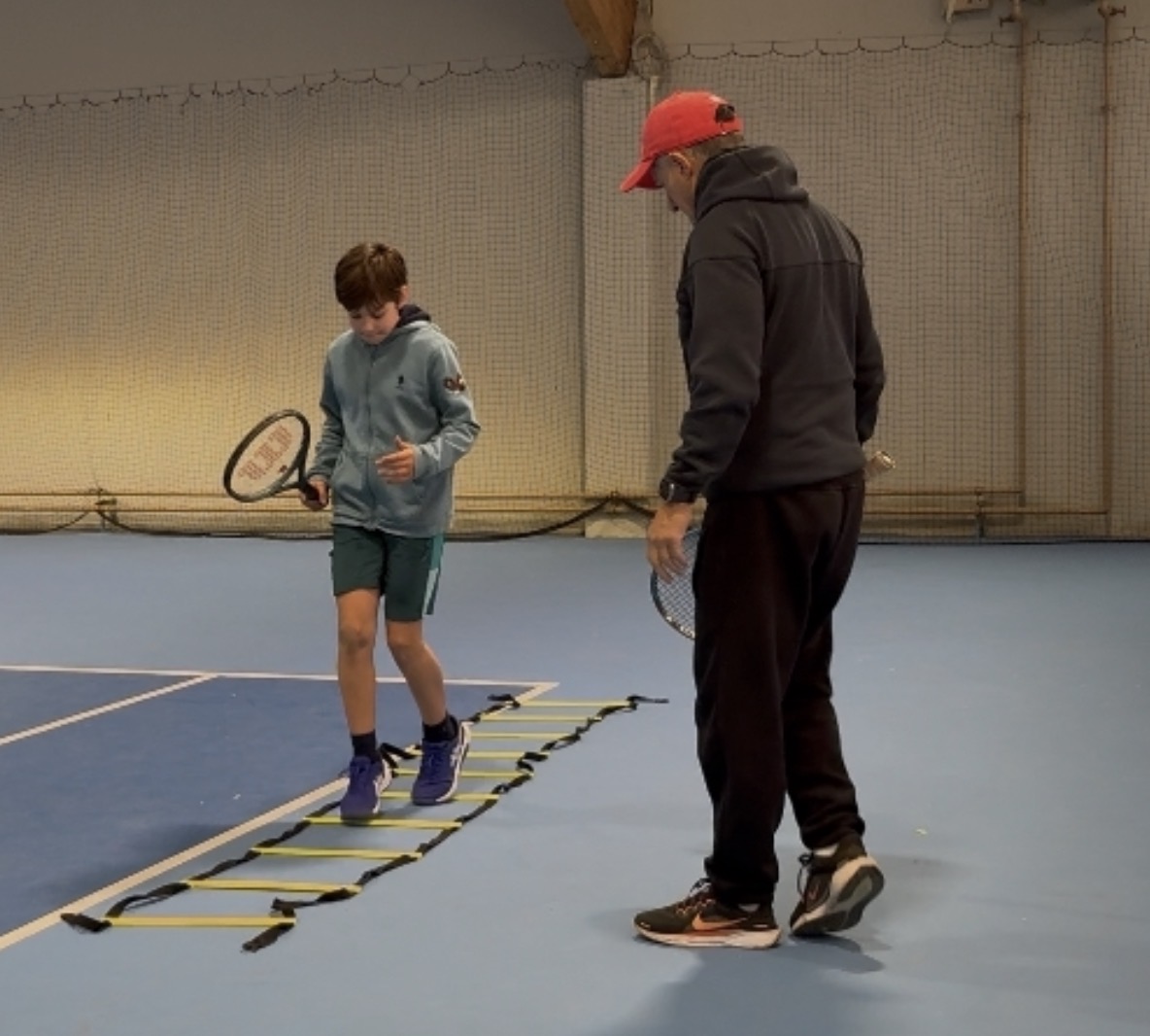 Trainer mit roter Kappe leitet Koordinationsleiter-Übung mit Kind in der Halle