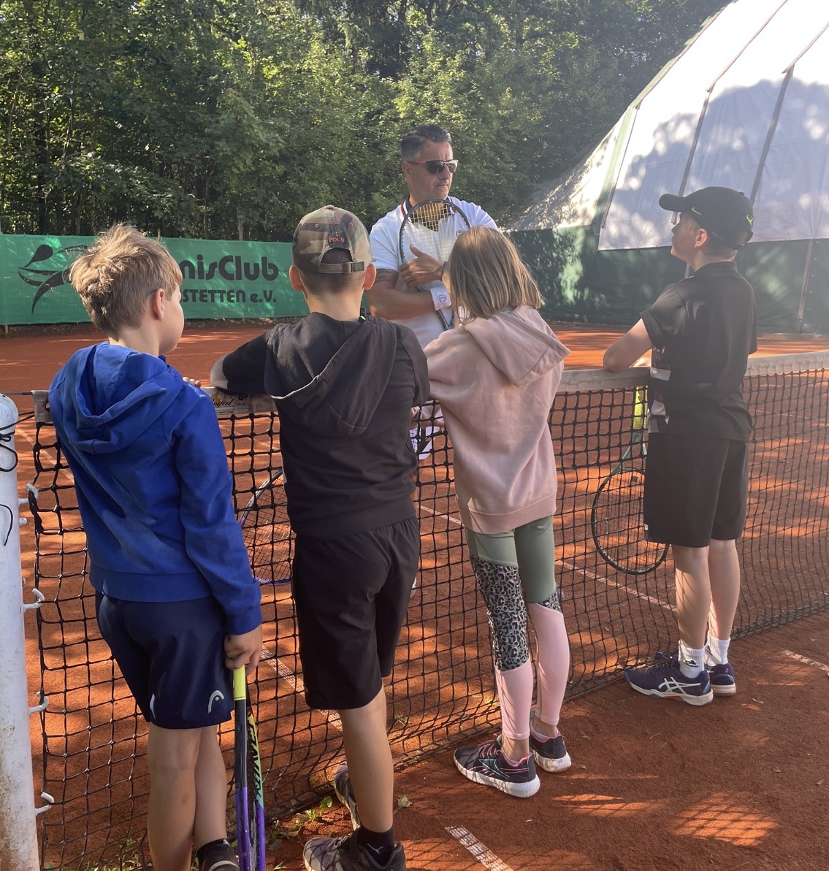 Trainer erklärt Kindern am Netz auf Sandplatz im TennisClub