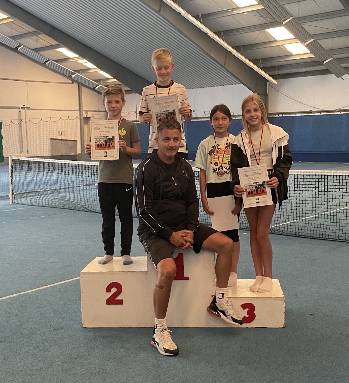 Trainer mit Kindern auf dem Siegerpodest mit Urkunden in der Tennishalle