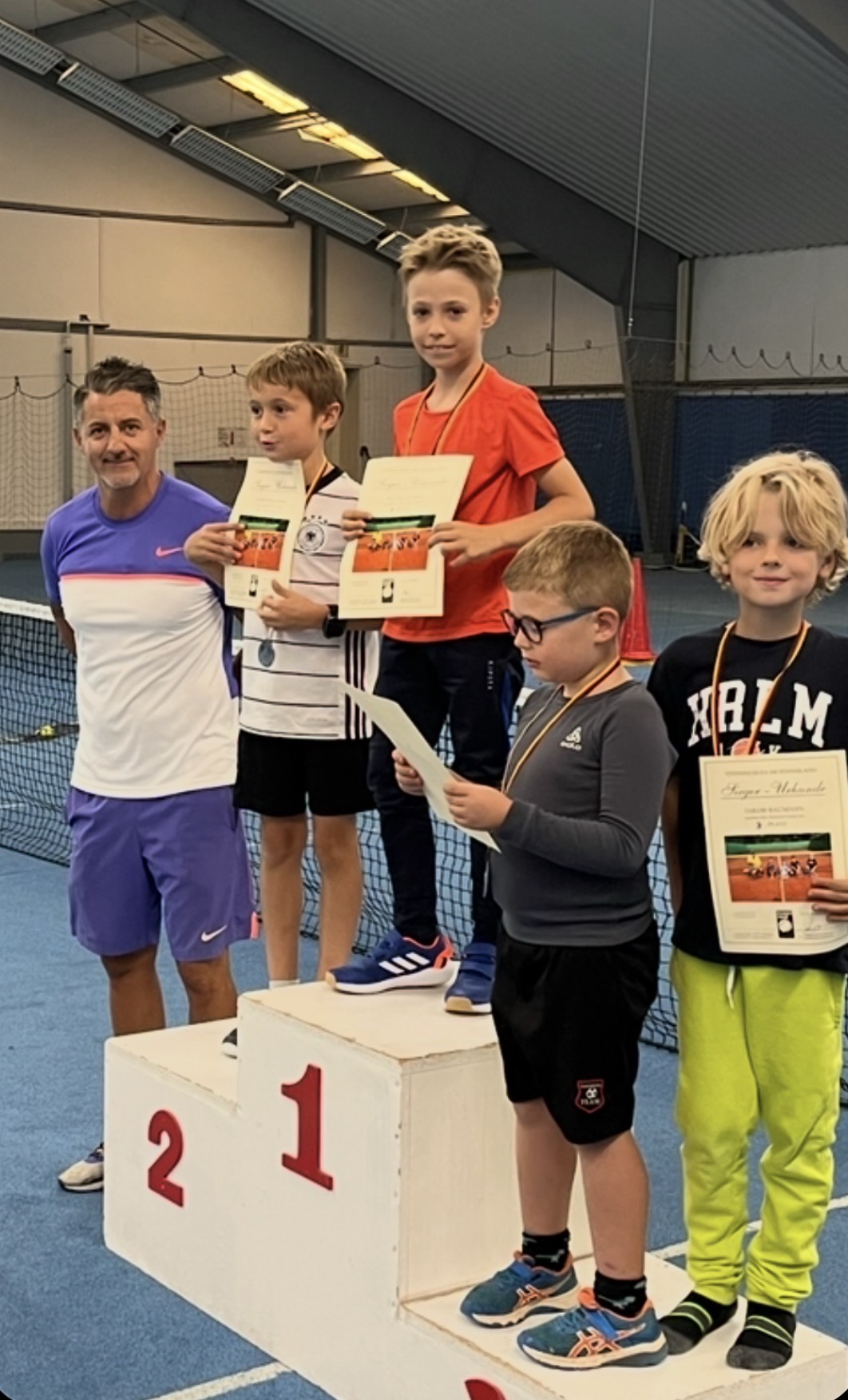 Trainer mit Kindern auf dem Siegerpodest mit Urkunden in der Tennishalle
