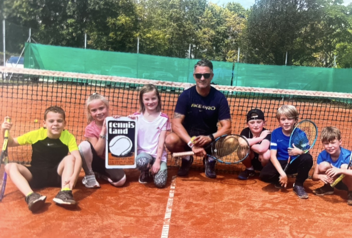 Trainer mit Kindergruppe und Tennisland-Schild am Netz auf Sandplatz