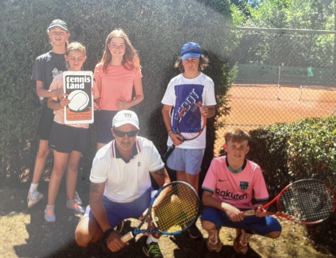 Trainer mit Kindergruppe und Tennisland-Schild vor Hecke auf Sandplatz